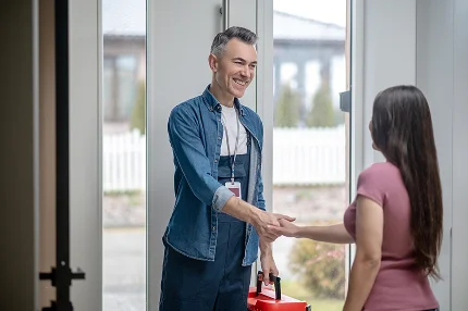 A bejáratnál egy farmeringben, szerszámosládát tartó férfi mosolyogva kezet ráz egy nővel. A jelenet barátságos és professzionális hangulatot áraszt.
