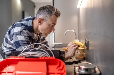 Kockás ingben lévő férfi, aki a konyhában javít egy fali aljzatot, koncentrált arckifejezéssel. Előtérben egy piros szerszámosláda vezetékekkel, a háttérben elmosódott konyhai eszközök.